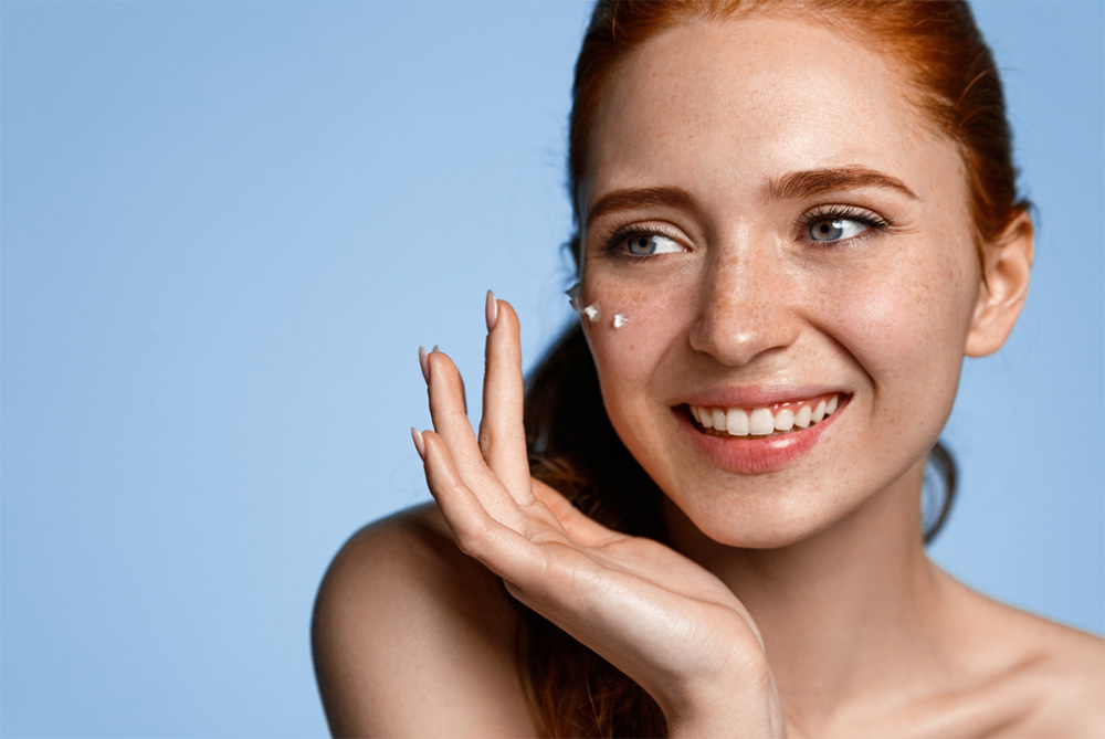 blue background with a red head applying dots of moisturizer to her freckled skin with a smile